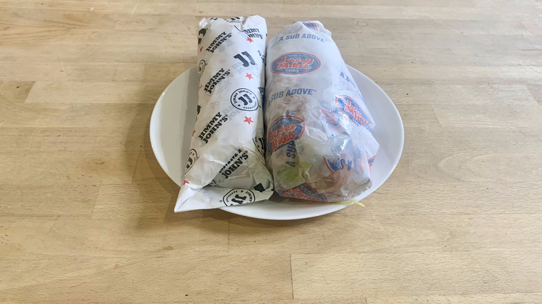 Jimmy John's and Jersey Mike's wrapped italian subs on white plate with wood background