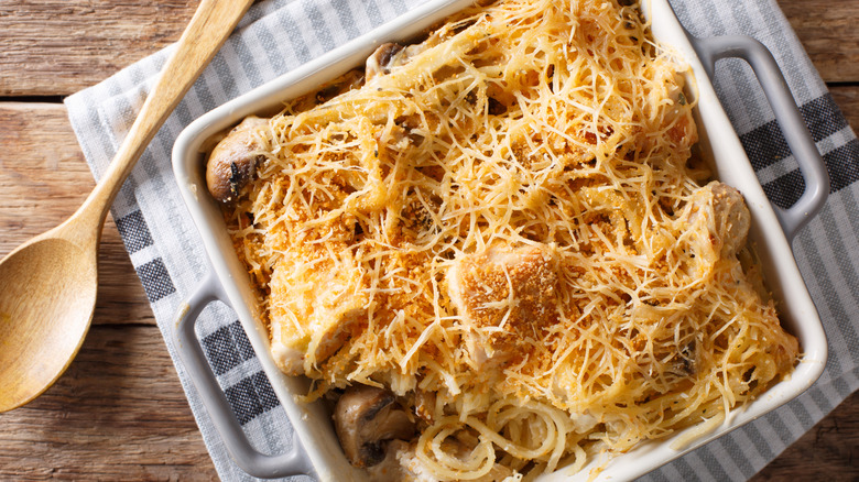 Baked chicken spaghetti in a white pan on top of a striped tea towel with a wooden spoon nearby.