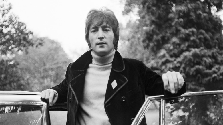 A black and white photo of John Lennon standing beside a car with trees in the background