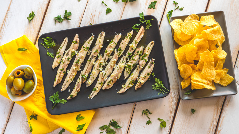 dishes of olives, white anchovies, and potato chips on a white wood table with fresh herbs