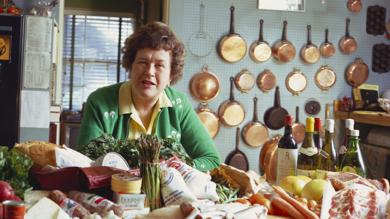 Julia Child behind a counter full of ingredients and a wall of copper pots behind her