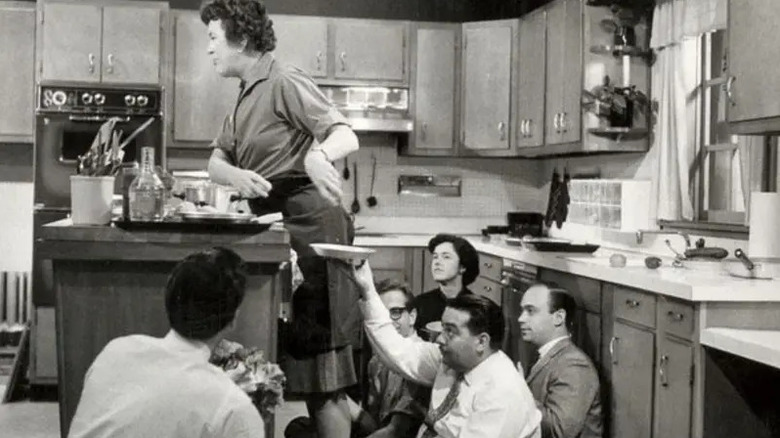 Julia Child stands during the taping of an episode of "The French Chef", with multiple crew members sitting out of sight of the camera, with one holding up a plate