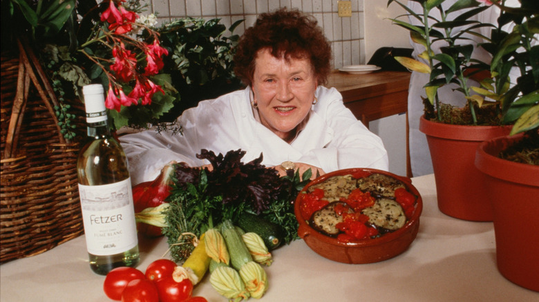 Julia Child sits in front of a table of fresh vegetables, an eggplant dish, and a bottle of white wine.