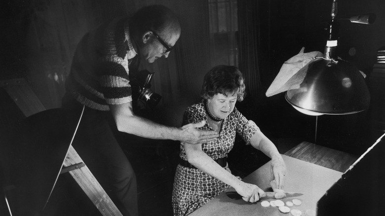 Julia Child chops a vegetable while her husband Paul sets up a photograph.