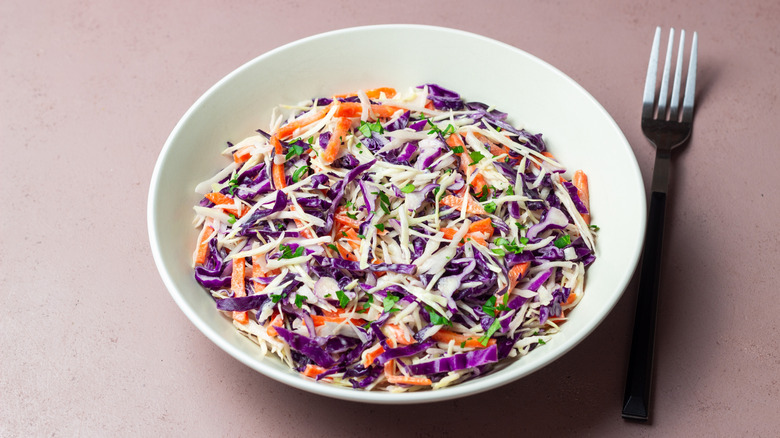 A bowl of classic coleslaw on a pink surface with a fork next to it.