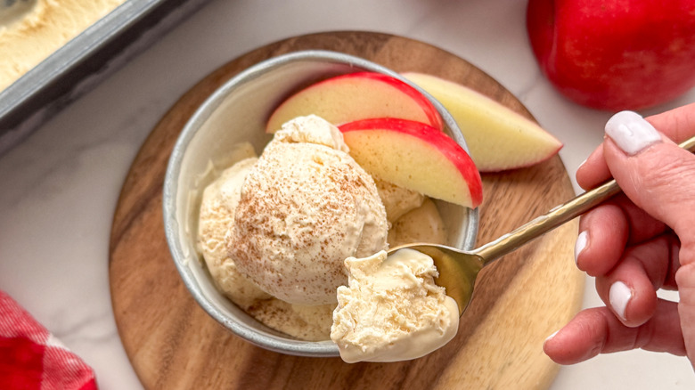 person holding a spoonful of apple cider ice cream above a bowl of ice cream and sliced apples