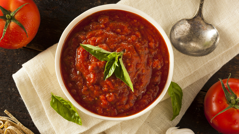 A bowl full of freshly prepared marinara sauce, with a ladle, tomatoes, and basil leaves nearby