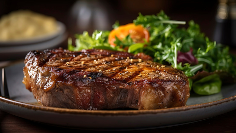 Close up of a grilled ribeye steak on a plate with a side salad in the background