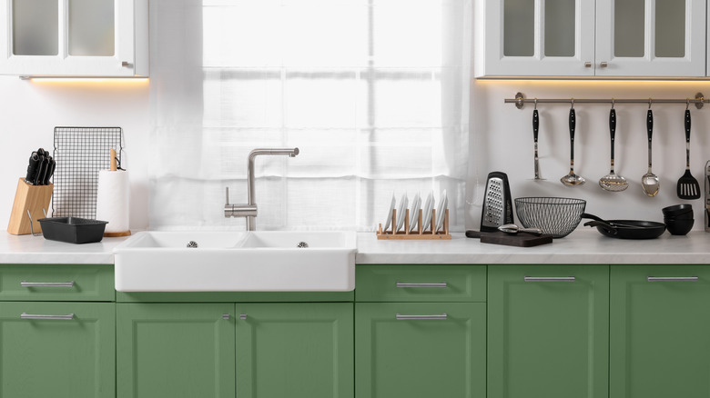 Sage green kitchen cabinets with below white countertop and sink with utensils out
