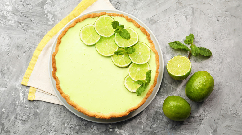 Whole key lime pie garnished with lime slices next to whole and halved limes on gray surface