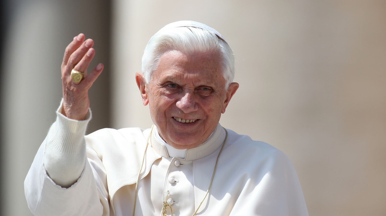 Pope Benedict XVI in white robes smiling and raising his hand in a blessing