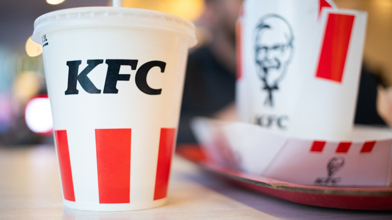 A KFC drink cup with some branded KFC cardboard food containers out of focus in the background