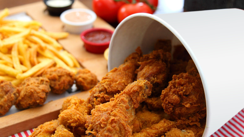 A upturned bucket of fried chicken in the foreground, with fries and dipping sauces in the background