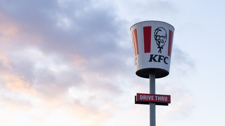 A giant KFC bucket sign on a pole that says drive-thru against a pale, cloudy sky