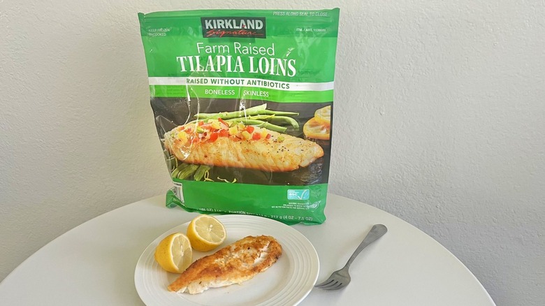 A cooked Kirkland Signature tilapia loin with a halved lemon on a plate with a bag of Kirkland Farm Raised tilapia loins behind