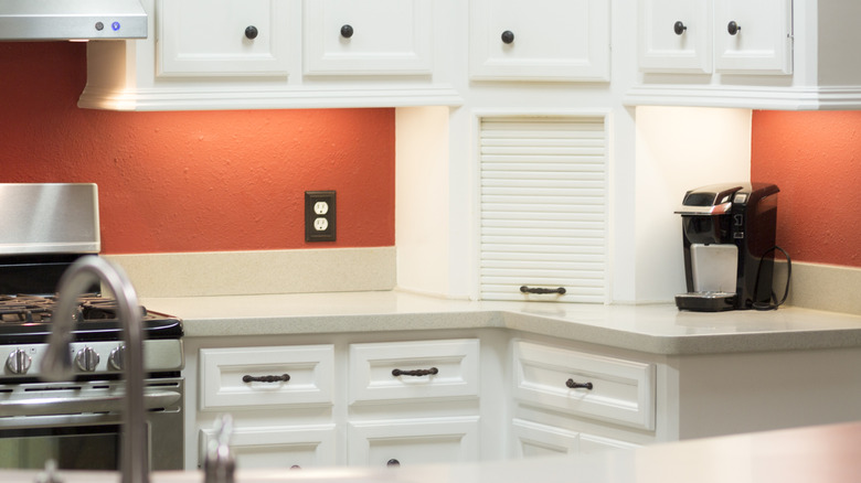 A white appliance garage sits in the corner of a kitchen counter in front of a red-orange backsplash.
