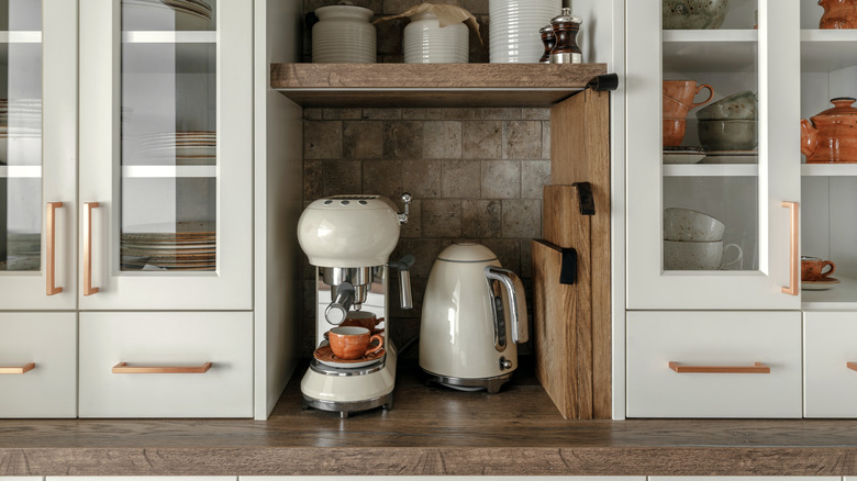 A coffee maker and a kettle sit in a nook in a stylish off-white kitchen with wood countertops.