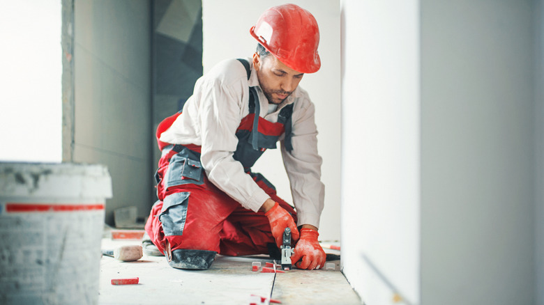 tradesman in red hard hat, denim coveralls, and collared shirt, installing tile flooring