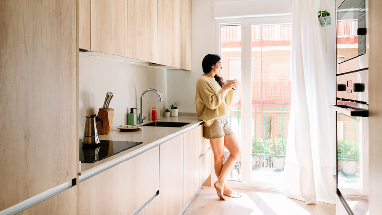 A woman stands with a mug looking out her window
