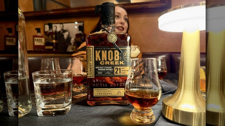 A bottle and glass of Knob Creek 21 Year bourbon on a table next to a glass of water and a lamp