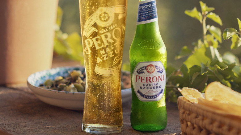A bottle of Peroni Nastro Azzurro on a table next to a filled glass, surrounded by food and plants