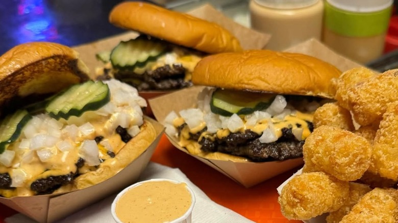 Three smashed burgers from Stay Tuned Burgers with tater tots and small cup of sauce on the side