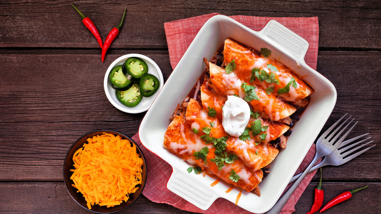 Enchiladas rojas in a baking dish next to forks, peppers, and bowls of cheese and jalapenos