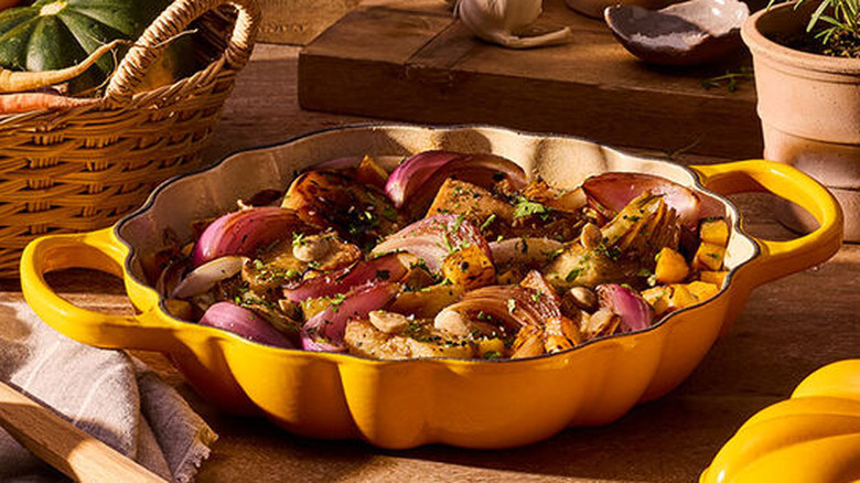 The Signature Pumpkin Braiser with the lid off filled with colorful roasted vegetables with a basket of veggies, bulbs of garlic, onions, and two pots of herbs.