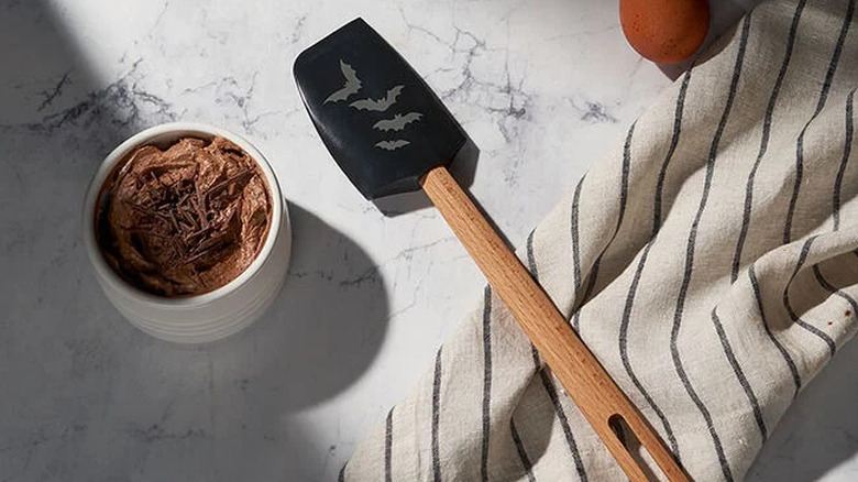 The Signature Spatula with Bat Appliqué lying on a counter on top of a white tea towel with black stripes and next to a white ramekin of chocolate mousse topped with chocolate shavings.
