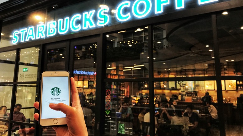 A hand holding an iPhone showing the Starbucks app in front of a Starbucks