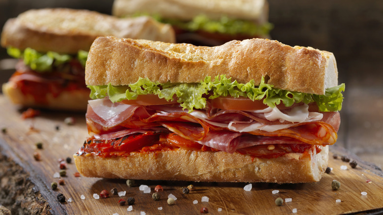 A stacked Italian sub with deli meats, lettuce and tomato on a baguette surrounded by salt flakes and peppercorns.