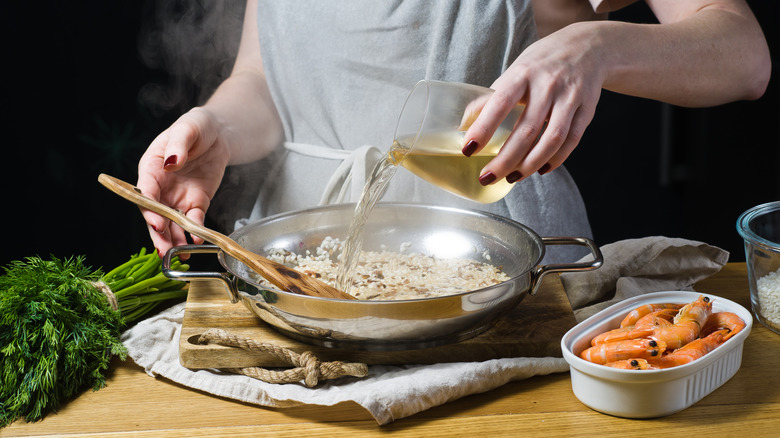 Cook pouring white wine into a pan of risotto surrounded by green herbs and shrimps