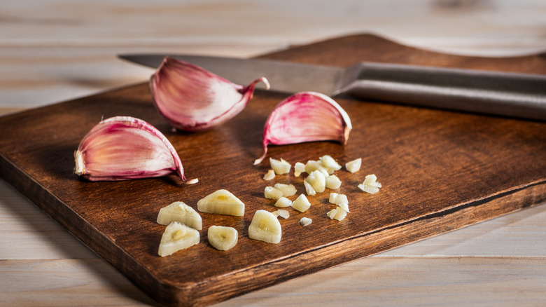 Whole and sliced garlic cloves on a dark brown cutting board with a sharp knife