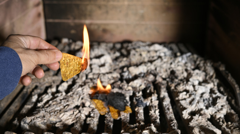 A hand holding a burning Dorito while a couple other Doritos smolder on an ash-covered grill in the background