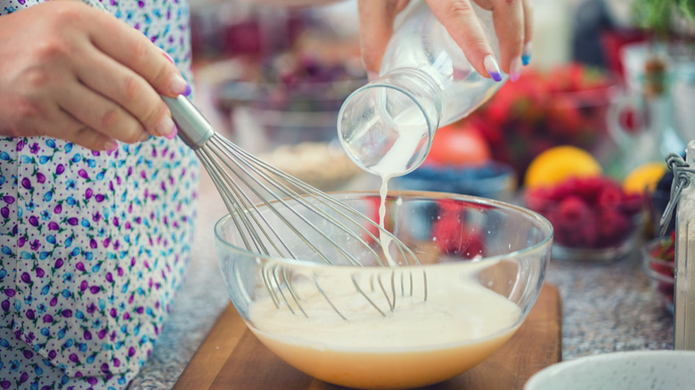 A partially visible person prepares a bowl of pancake batter, pouring milk with one hand and holding a whisk with the other
