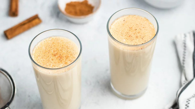 Two glasses of Creamy Coquito dusted with cinnamon with more cinnamon in the back