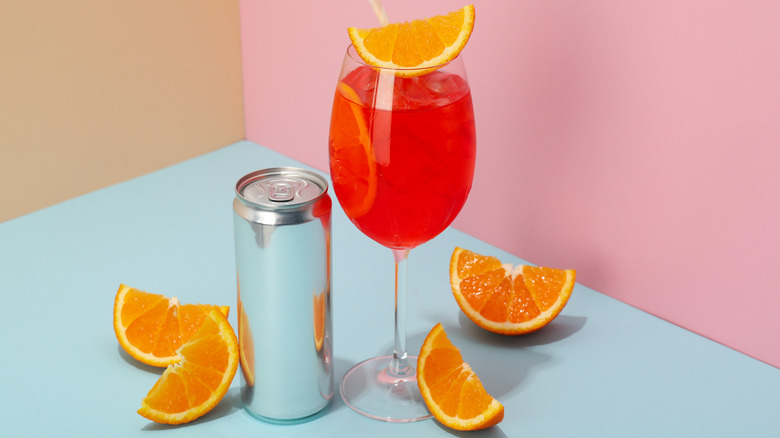 A canned cocktail with poured drink next to it in a glass, orange slices around them