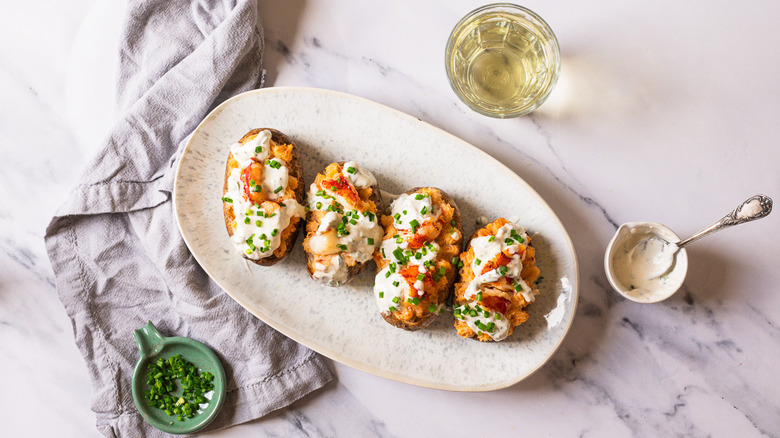Four lobster-stuffed baked potatoes on a platter with sauce, chives, and glass of wine.