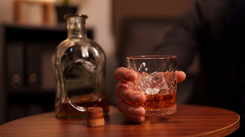 A hand holds a glass of whisky next to an open decanter.