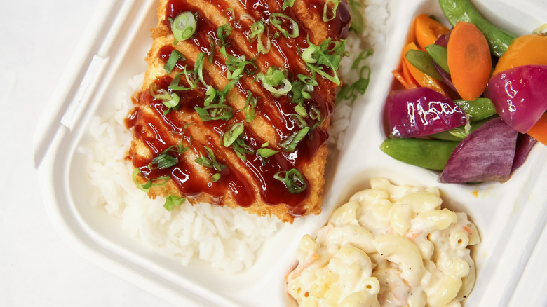 A Hawaiian plate lunch in a to-go container with macaroni salad, vegetables, rice, and fried fish topped with sauce and scallions