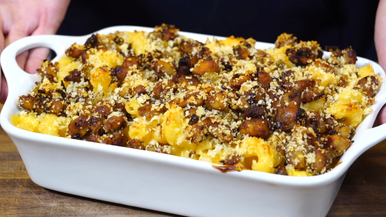 A casserole dish of baked mac and cheese topped with stuffing is lifted by an unseen person with both hands
