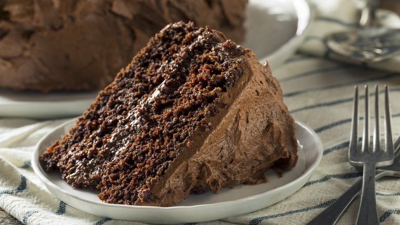 A large slice of chocolate cake on a white plate next to a fork, with a cake in the background