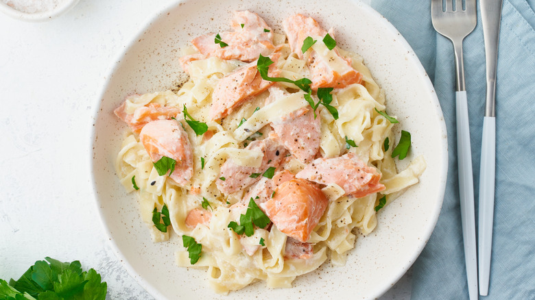 White plate of fettuccine pasta with chunks of salmon, creamy sauce, and fresh green herb garnish.
