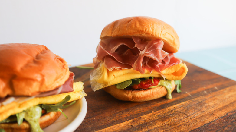 a prosciutto egg and pesto breakfast sandwich sits on a wooden plank
