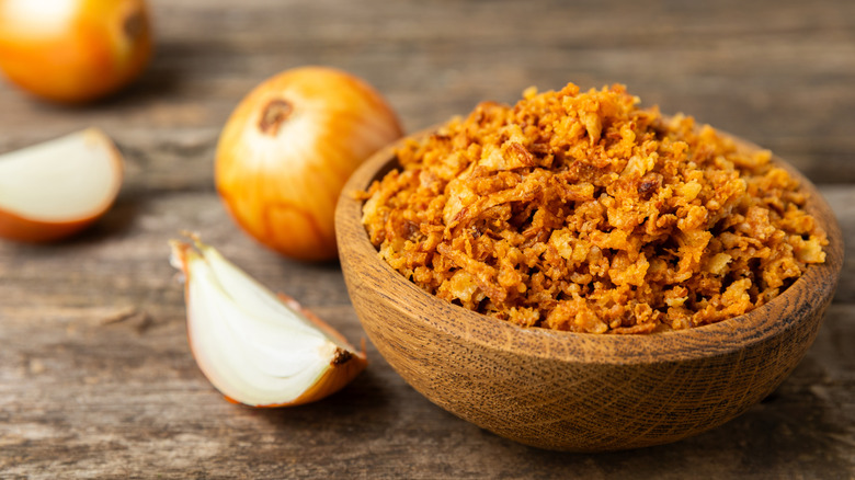 A wooden bowl of crispy fried onions next to some raw onions on a wooden table