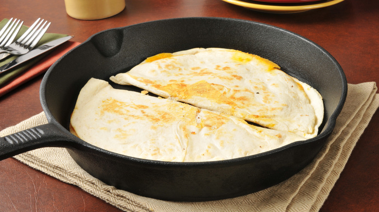 Two quesadillas in a large cast iron skillet sitting atop a tea towel on a table with plates and cutlery