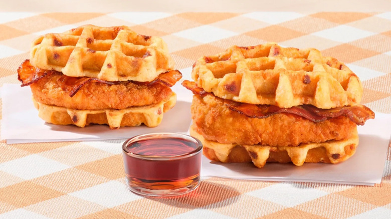 two Chick-Fil-A chicken and waffles breakfast sandwiches sitting on napkins atop a checkered tablecloth with a small ramekin of syrup