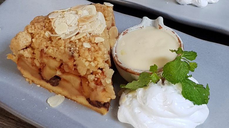 A slice of apple pie on a plate with cream, custard, and mint leaves.
