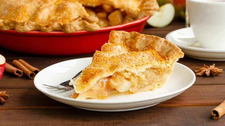 A slice of golden, sugar-topped apple pie surrounded by cinnamon sticks with a coffee cup and the full pie in the background.