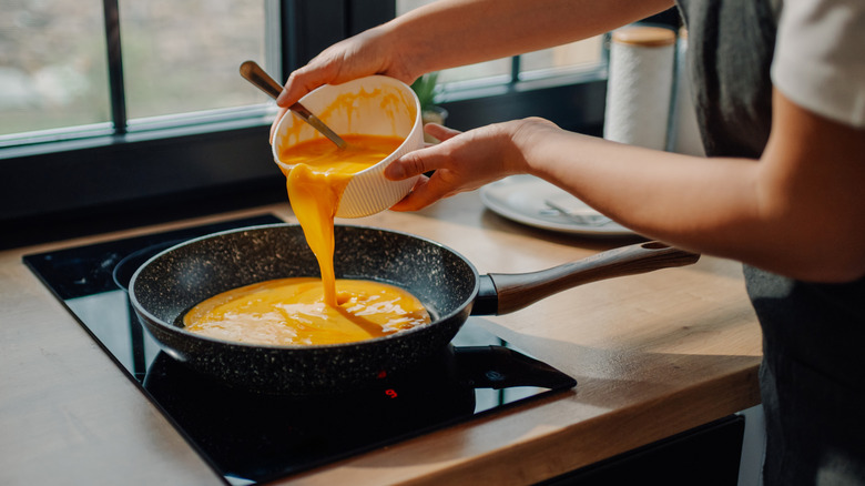 a cook starting an omelet by pouring whisked eggs into a skillet on the stove